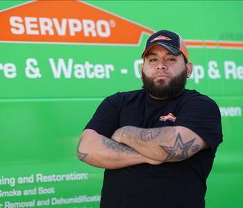 A man with black hair and a beard wearing a logo ball cap and T-shirt, standing with arms folded next to a SERVPRO truck