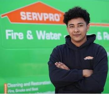 Young, clean-shaven man with mid-length, dark, curly hair wearing a black SERVPRO hoodie, standing next to a SERVPRO truck