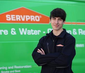 Tall young man with bushy dark hair wearing a SERVPRO logo jacket, standing with arms folded at the side of a SERVPRO truck