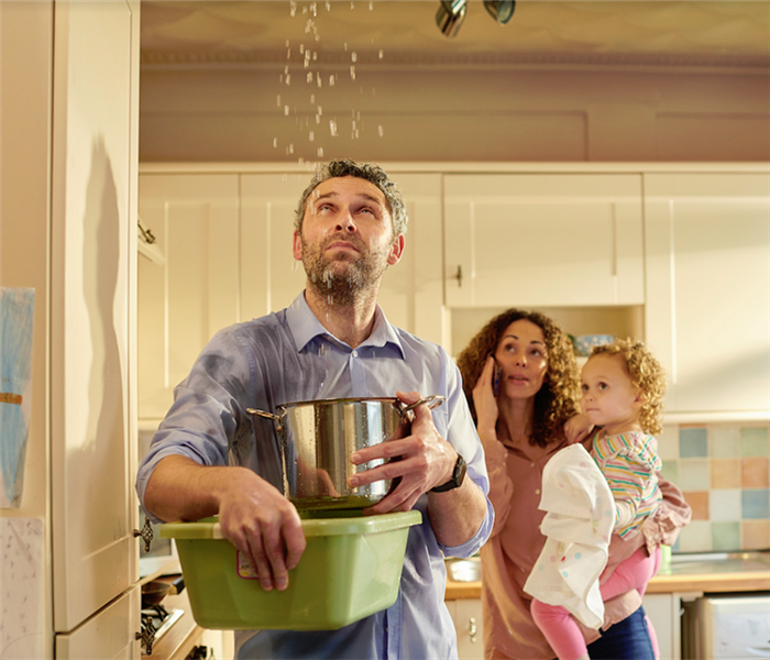 a man holding a bucket to catch water falling from his ceiling while his wife is on the phone in the background