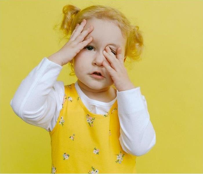 Toddler in white,long sleeve shirt, yellow jumper standing in front of yellow wall. Child has hands on face, looking stressed