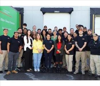 The SERVPRO of Gaithersburg/Germantown Team, in SERVPRO uniforms, standing in 3 rows facing front, with SERVPRO trucks behind