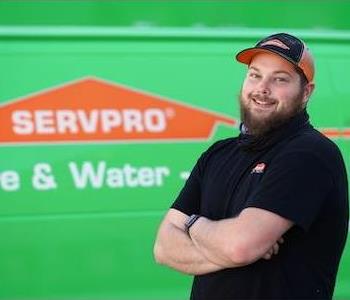 Tall young man with reddish hair and a long beard wearing a SERVPRO polo and ball cap with a friendly smile by SERVPRO truck