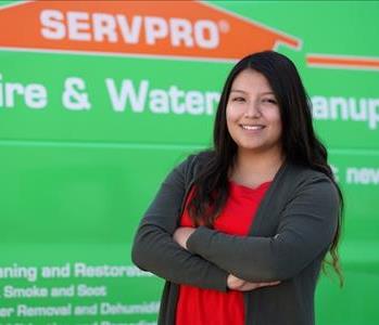 Young woman with long dark hair smiling with arms folded standing next to a SERVPRO truck